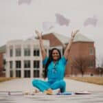 doctor in blue scrubs throwing notes in the air sitting on the walkway in front of the hospital among books